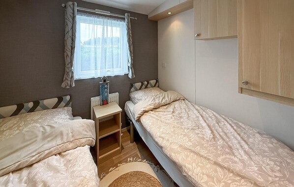 A bedroom with two single beds, a window with curtains, a radiator, and a small wooden shelf.