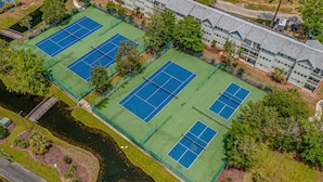 Ilmakuva tenniskenttäalueesta, jossa on useita kenttiä, ja aluetta ympäröivät puut ja rakennus.