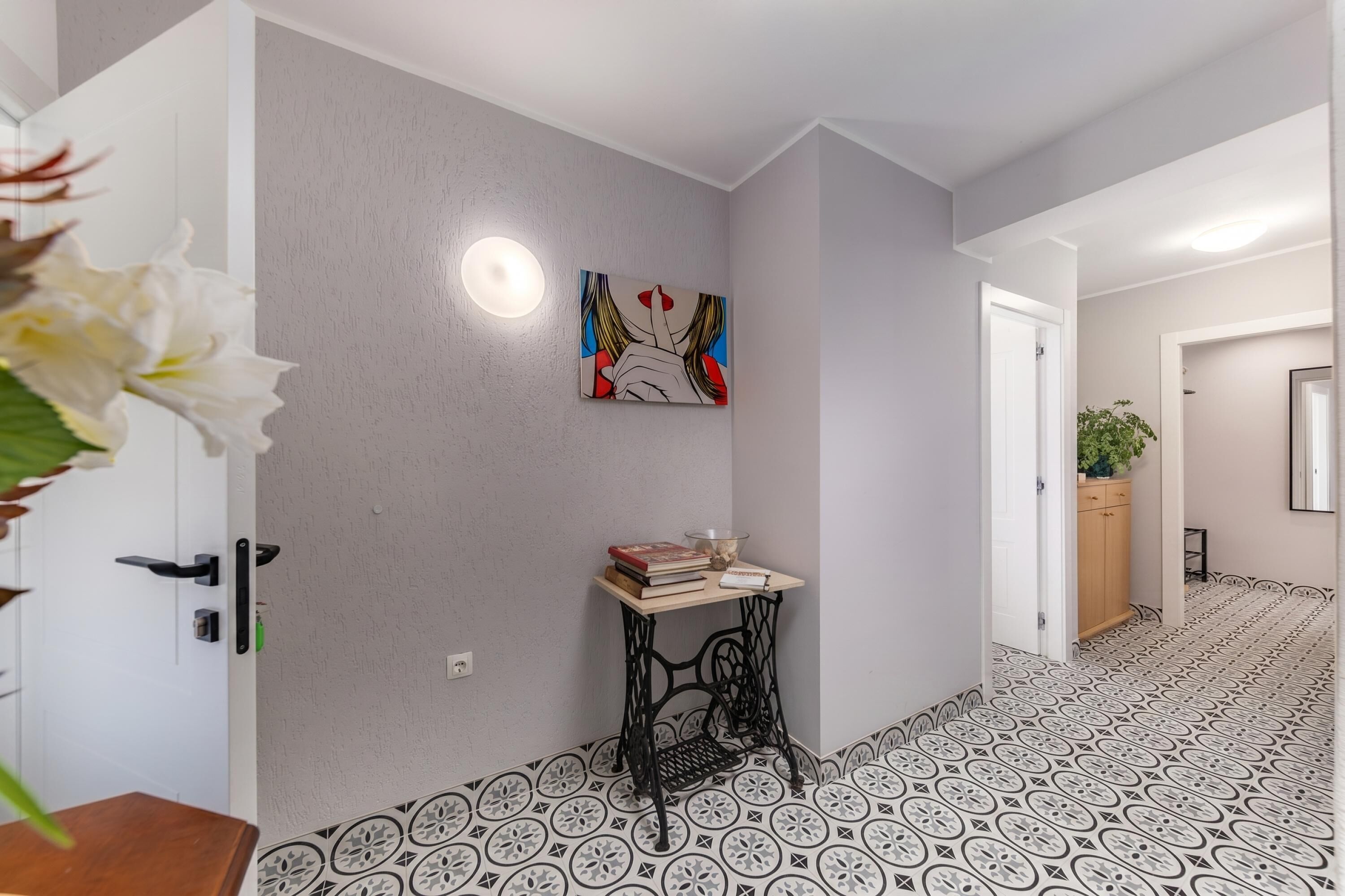 A hallway with patterned flooring, a table with books and a bowl, a painting on the wall, and a door with a light fixture.