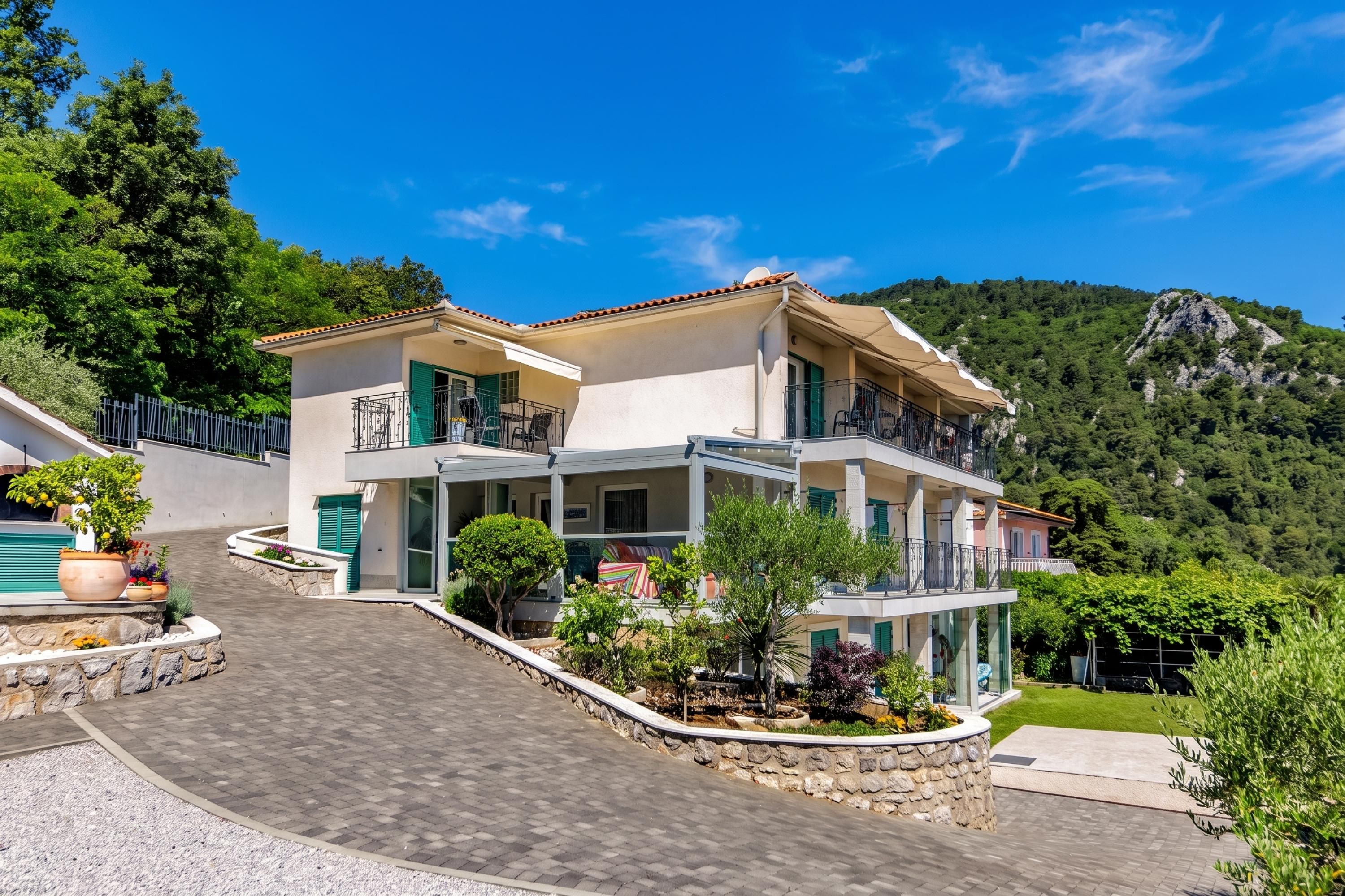 A two-story house with a balcony, surrounded by greenery and a mountain backdrop.