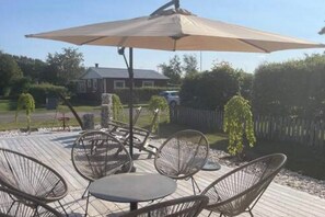 En udendørs terrasse med metalstole og et bord, en stor parasol og et hus i baggrunden.