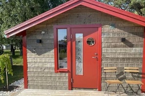 Een klein houten huisje met een rode deur, een raam en twee houten bankjes buiten.