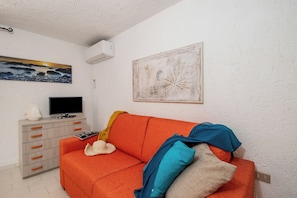 A room with a red sofa, a television on a stand, a white dresser, and a wall-mounted air conditioner. - Ladunia 2, Marinella, Italy (Marinella)