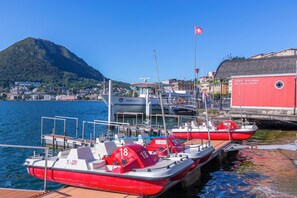 Un puerto deportivo con varias embarcaciones rojas y blancas amarradas, un edificio rojo con señalización y un paisaje montañoso de fondo.
