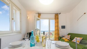 Un coin repas avec une table dressée pour deux personnes, une vue sur la mer par la fenêtre et un canapé vert agrémenté de coussins à motifs.