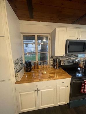 A kitchen with white cabinets, a wooden countertop, a microwave, a stove, and a window with a view of a house and greenery. - Hotel Style Lakeview Cottage • 3BR • Heated Pool (Lake Wales)