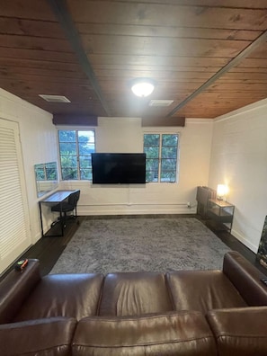 A room with a large flat-screen TV mounted on the wall, a brown leather sofa, a small desk with a chair, and a window with a view of greenery.