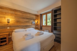 Ein Schlafzimmer mit hölzernem Kopfteil, einem Bett mit weißen Bettwäsche, zwei Wandleuchten, einem Fenster mit Vorhängen und einer eingebauten Regaleinheit.
