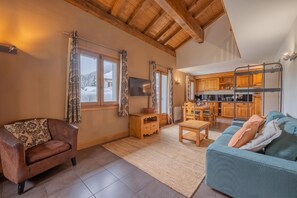 A cozy living room with a brown leather sofa, a blue sofa, and a wooden coffee table. There is a kitchen in the background with wooden cabinets and a dining area.