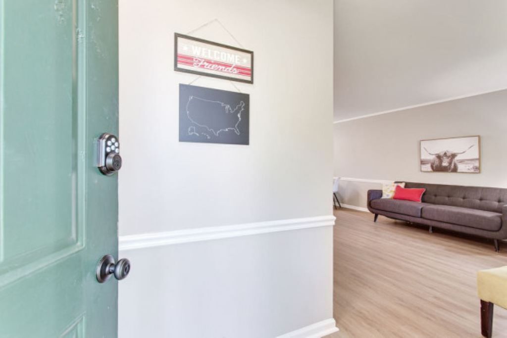 A room with a green door, a white wall, a framed welcome sign, a map, a sofa with a red pillow, and a framed picture of a bull.