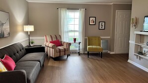 Un salón con un sofá de color oscuro, un sillón a rayas, una silla amarilla, una mesita con una lámpara y una ventana con cortinas.