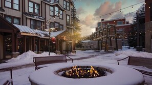 Un espacio al aire libre cubierto de nieve, con un brasero, bancos y edificios al fondo.