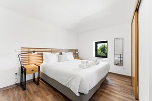 A modern bedroom with a wooden headboard, a bedside lamp, a small table, a large bed with white bedding, a window with a view of greenery, and a mirror on the wall. - GuestReady - Courtyar 9/99 - Apt 05 (Porto)