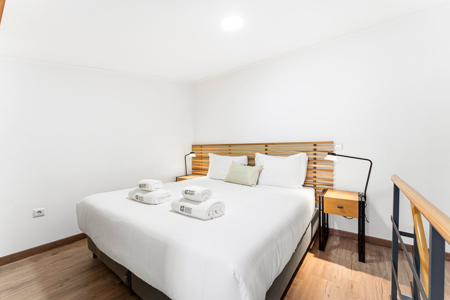 Hotelkamer met een bed, houten hoofdeinde, wit beddengoed, een nachtkastje met lamp, en opgerolde handdoeken met de tekst 'GUEST READY'.