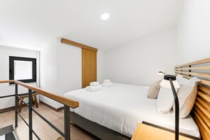 A modern hotel room with a bed, a wooden headboard, a nightstand, a desk, a window, and a door. - GuestReady - Courtyar 9/99 - Apt 7A (Porto)