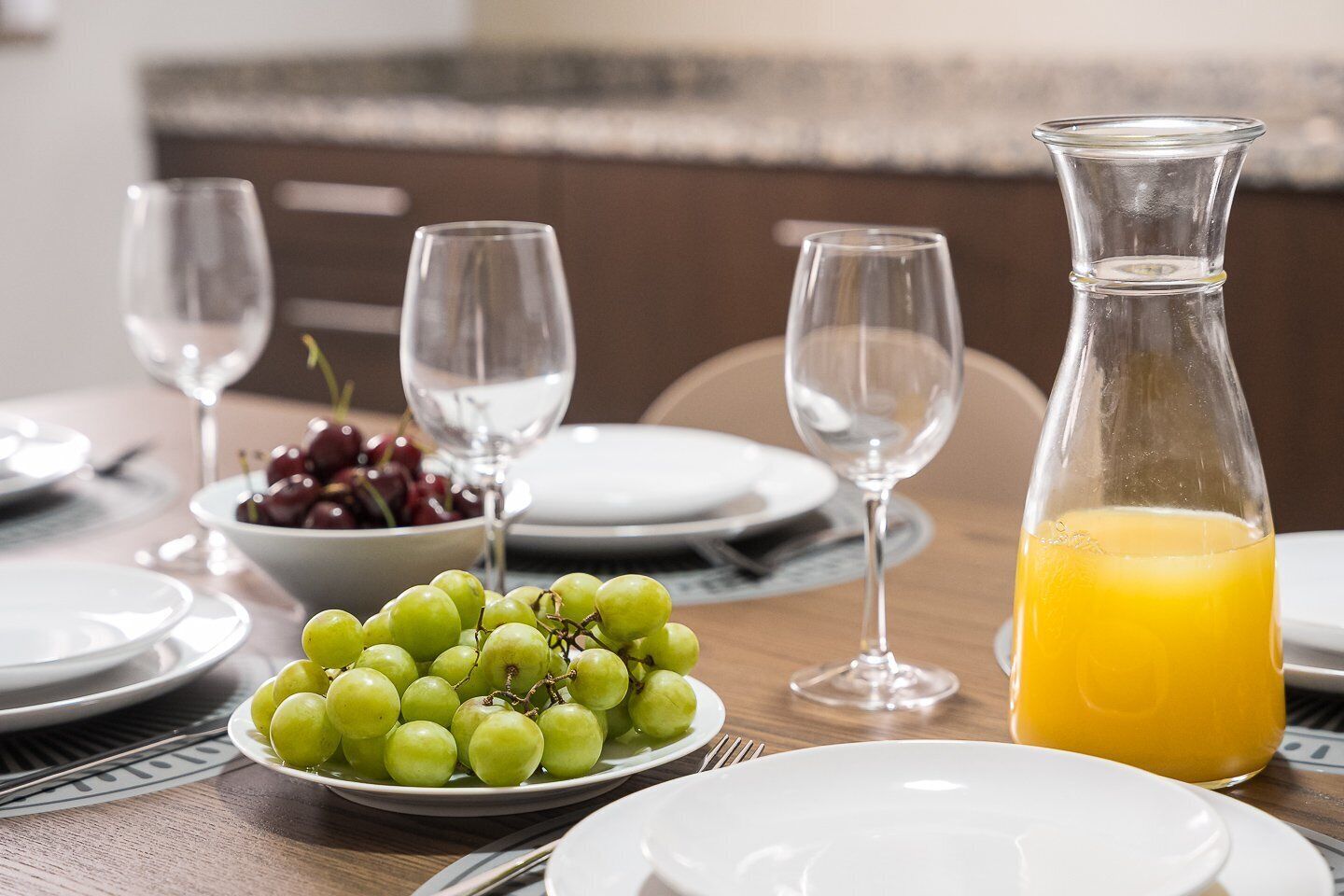 A dining table with plates, a bowl of grapes, a pitcher of orange juice, and empty wine glasses.