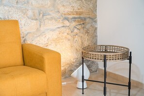 A room with a sofa, a wicker side table, and a stone wall. - GuestReady - Courtyar 9/99 - Apt 7A (Porto)