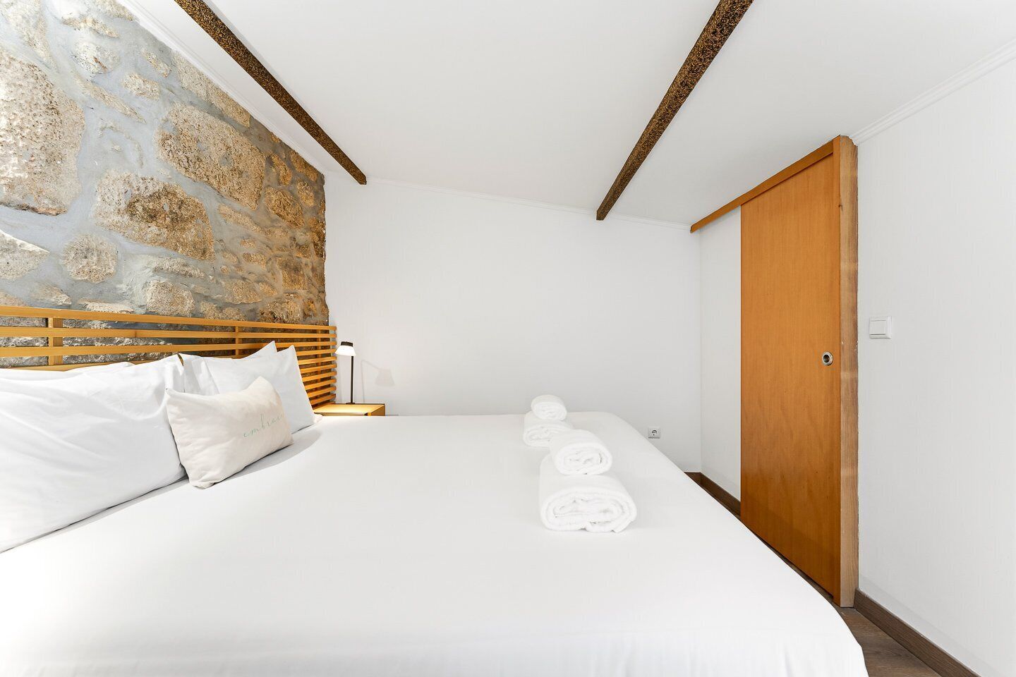 A bedroom with a bed, pillows, a nightstand, a lamp, a wooden headboard, a stone wall, and a wooden door.