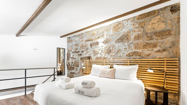Ein Hotelzimmer mit einer Wand aus Naturstein, einem Bett mit weißen Leinen, einem Holz-Kopfteil und einem Nachttisch mit einer Lampe.