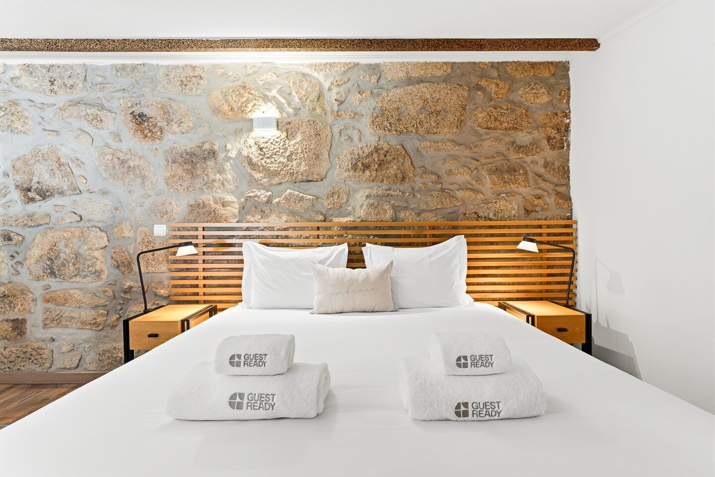 Quarto de hotel com parede de pedra, cabeceira de madeira, cama bem arrumada com roupa de cama branca e duas mesas de cabeceira com luminárias.