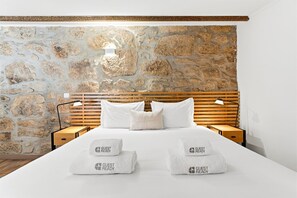 A hotel room with a stone wall, a wooden headboard, a neatly made bed with white linens, and two nightstands with lamps.