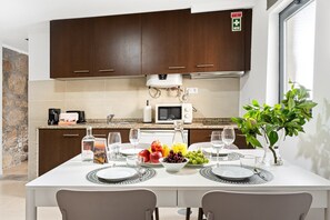 A modern kitchen with dark wooden cabinets, a white countertop, a microwave, and a dining table set for two with plates, glasses, and a fruit bowl.