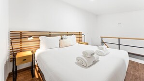 Una habitación de hotel con cabecera de madera, cama con sábanas blancas, dos mesitas de noche con lámparas y una toalla enrollada sobre la cama.