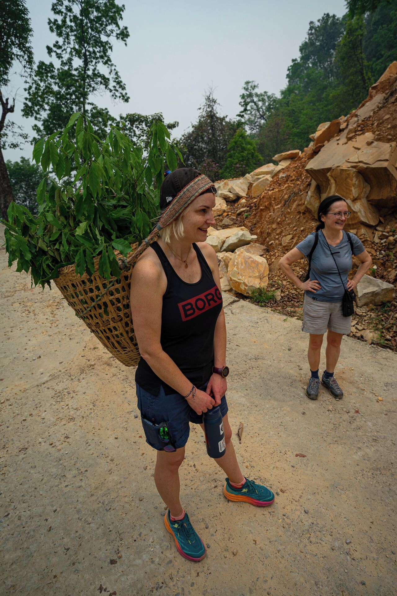 Two people standing on a dirt path with a rocky area and greenery in the background.