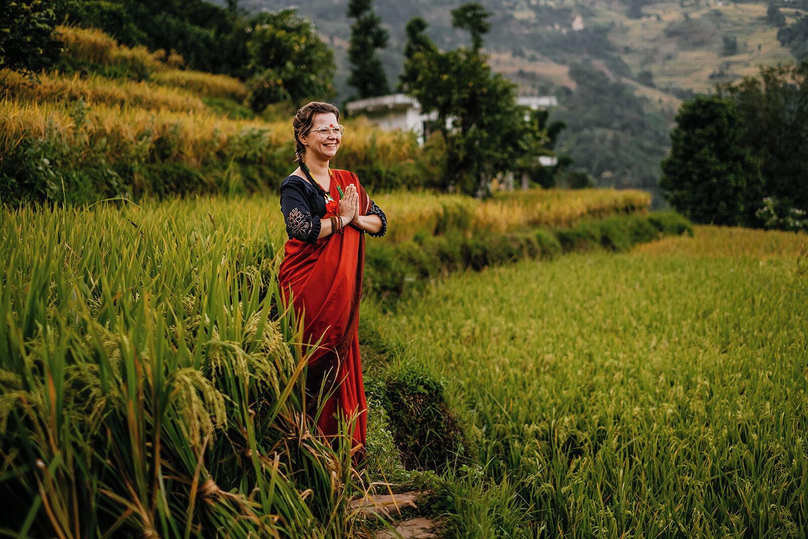 Una persona vestida con traje tradicional parada en un campo de arroz, con montañas al fondo.