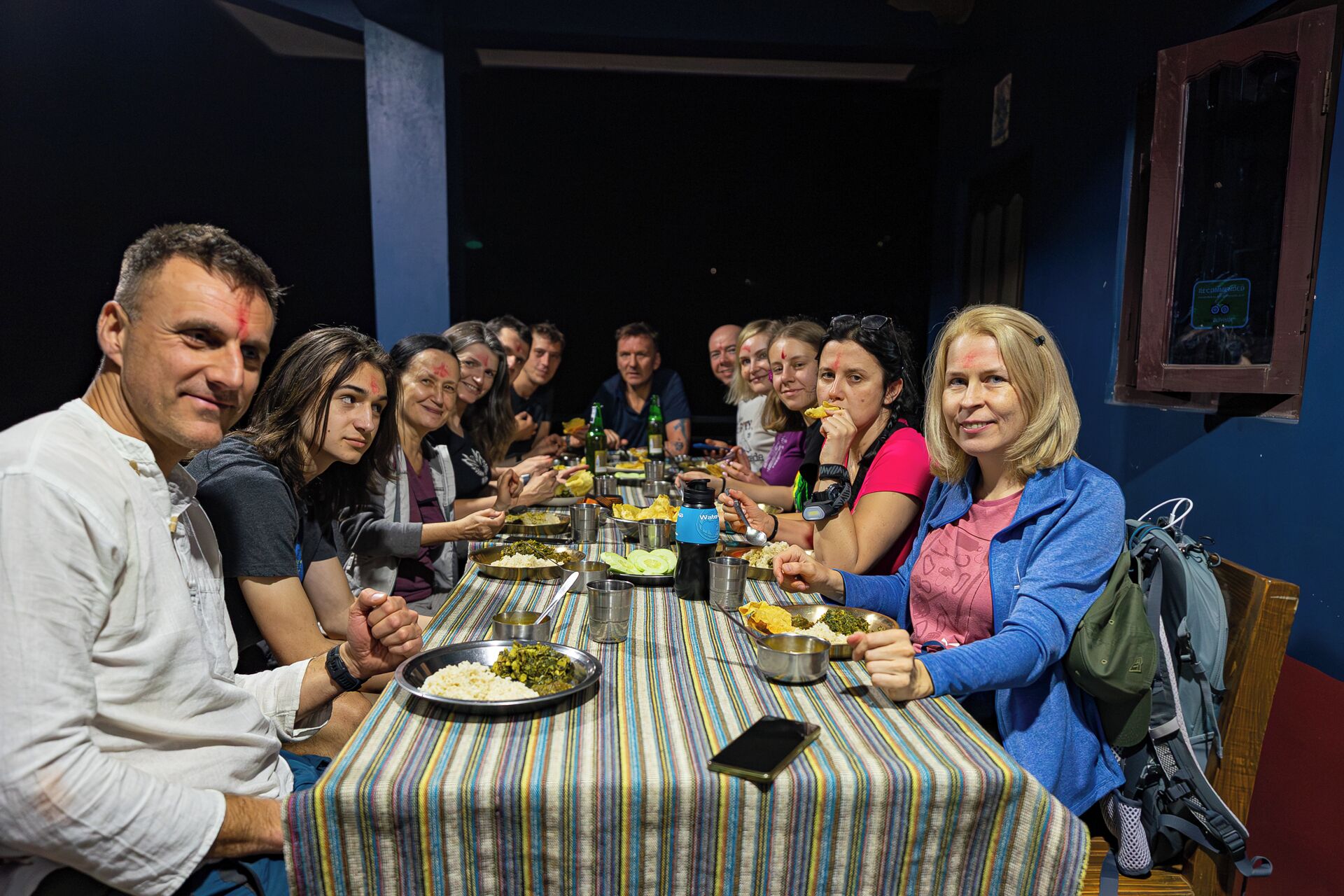 Un grupo de personas cenando en una mesa con platos de metal, un mantel a rayas y un teléfono inteligente sobre la mesa.