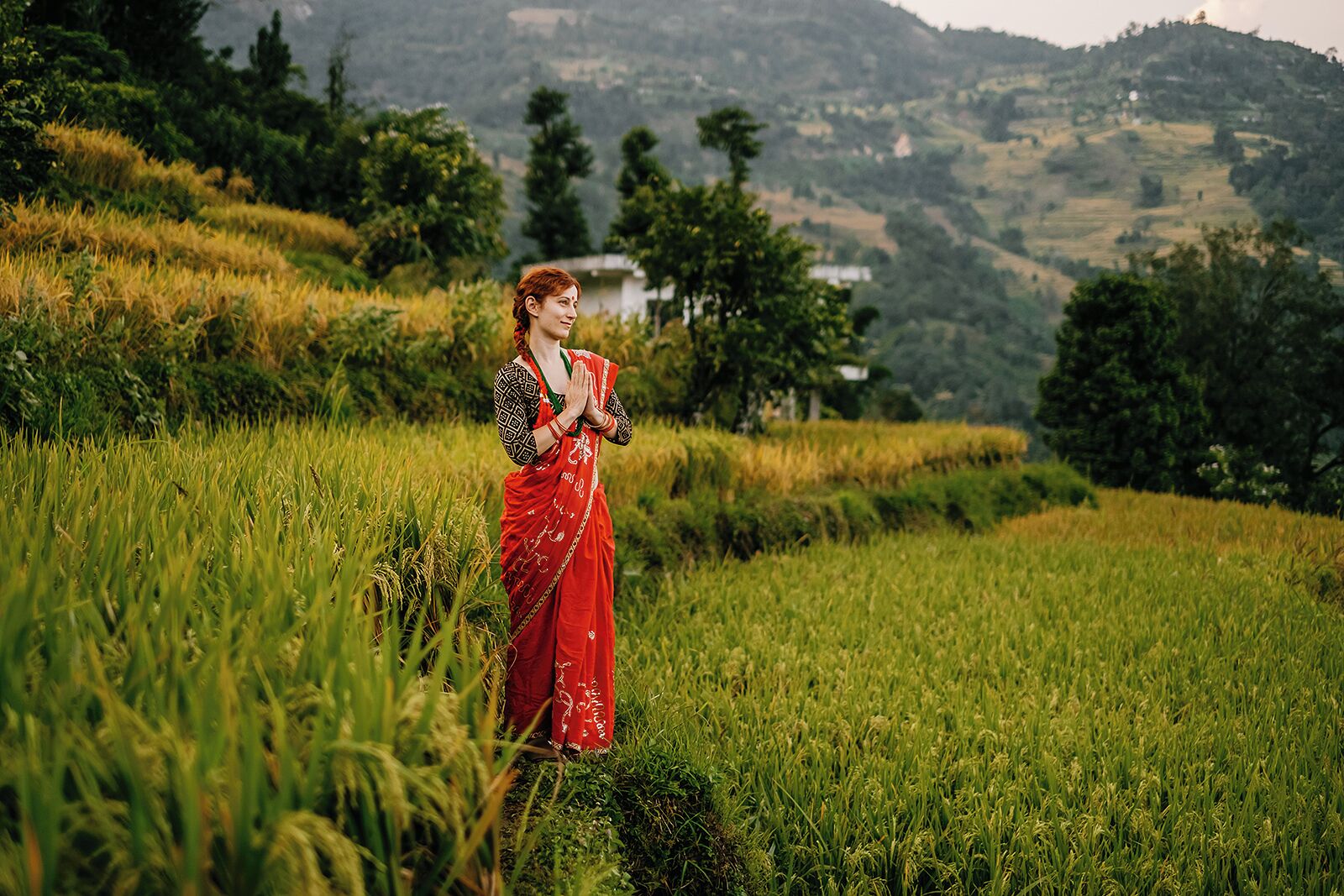 A woman in a red sari standing in a lush green field with a hilly landscape in the background.