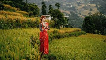 A woman in a red sari standing in a lush green field with a hilly landscape in the background.