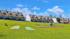 Una hilera de edificios residenciales con una pendiente cubierta de césped, un parque infantil y una cancha de tenis.