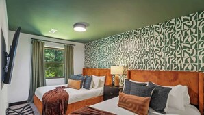 Habitación de hotel con dos camas, televisor, ventana con cortinas, pared con estampado y mesita de noche con lámpara.