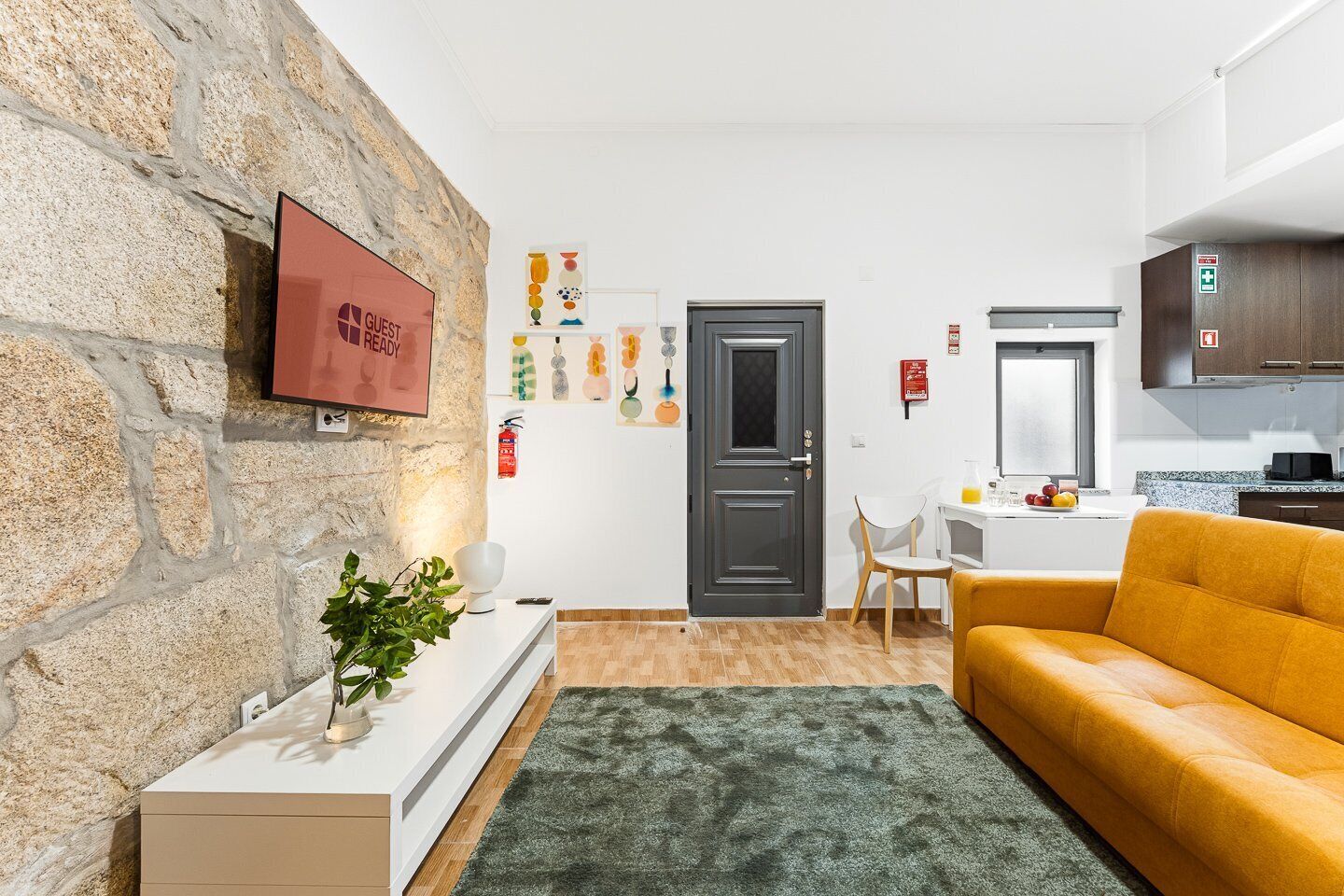 A modern living room with a stone wall, a flat-screen TV, a sofa, a coffee table, and a kitchen area with cabinets and a window.