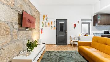 Ein modernes Wohnzimmer mit Steinwand, Flachbildfernseher, einer Couch, einem Couchtisch und einer Küchenzeile mit Schränken und einem Fenster.
