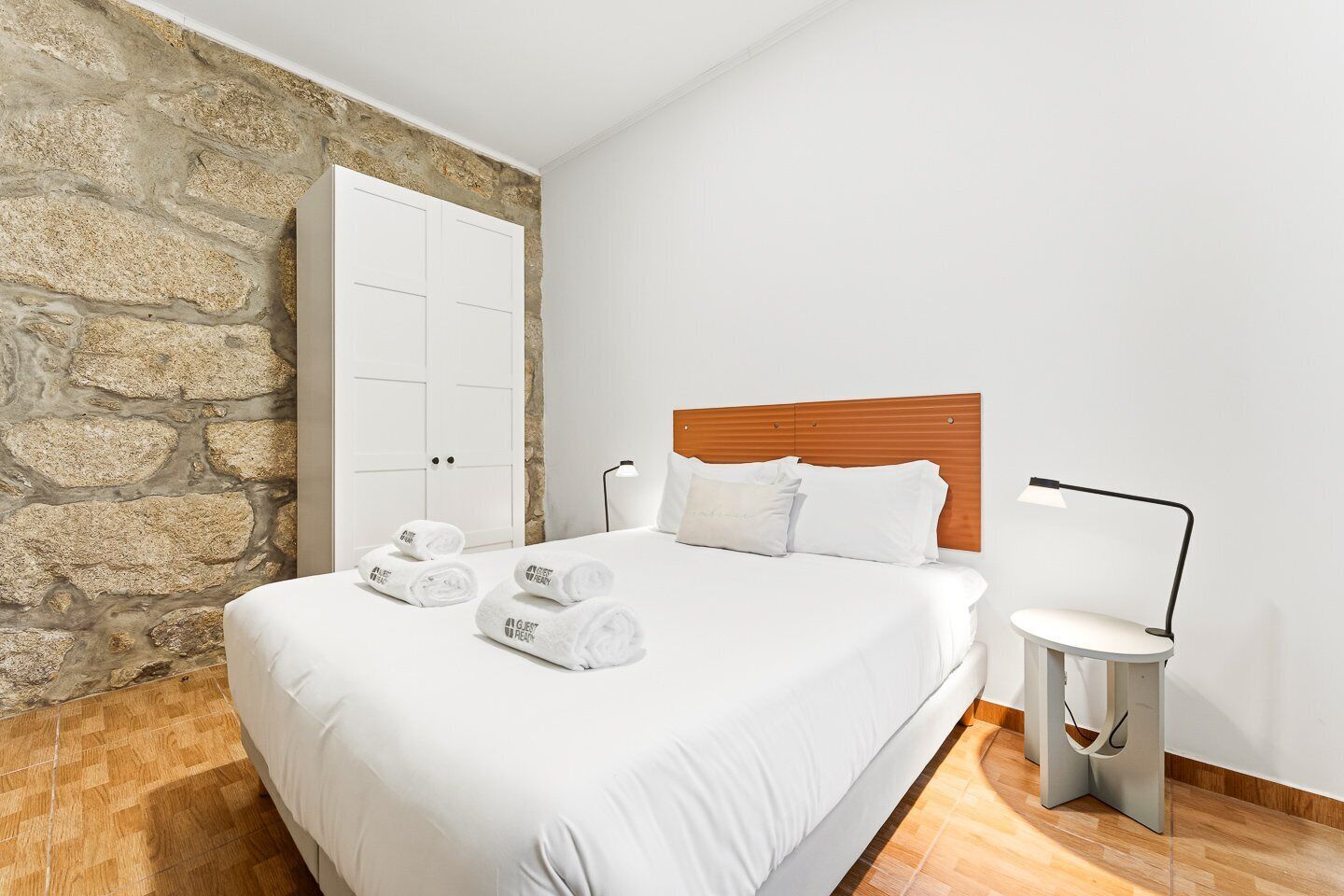 Ein Schlafzimmer mit Steinsturz, Holz Kopfteil, einem Bett mit weißen Leinen und einem Nachttisch mit Lampe. Auf dem Bett liegen zusammengerollte Handtücher.