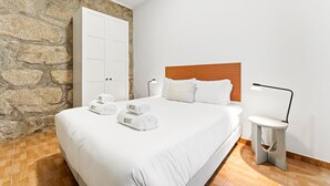 Ein Schlafzimmer mit Steinsturz, Holz Kopfteil, einem Bett mit weißen Leinen und einem Nachttisch mit Lampe. Auf dem Bett liegen zusammengerollte Handtücher.