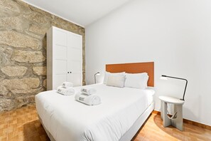 A bedroom with a stone wall, a wooden headboard, a bed with white linens, a bedside table with a lamp, and rolled towels on the bed. - GuestReady - Courtyar 9/99 - Apt 09 (Porto)