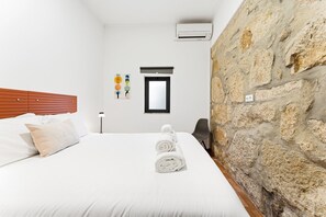 Ein Hotelzimmer mit Steinwand, einem Bett mit weißer Bettwäsche, einem Holzkopfende, einer Wand-Klimaanlage und einem kleinen Fenster.