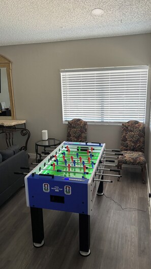 Una habitación con mesa de fútbol, sofá, sillón, ventana con persianas y espejo en la pared.