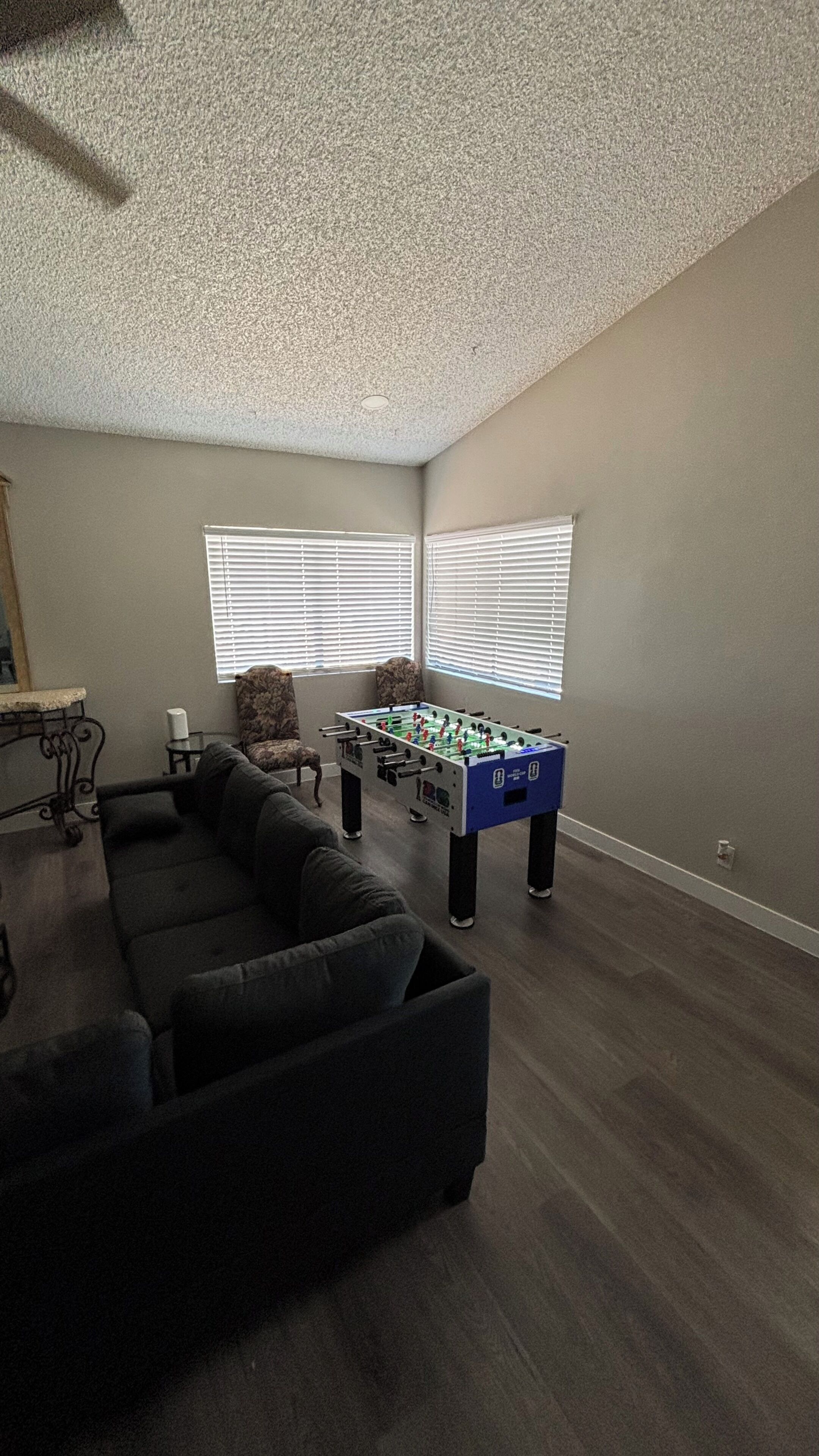 Una habitación con un sofá de color oscuro, una mesa de futbolín y una ventana con persianas.