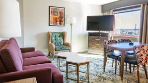 Une chambre d’hôtel équipée d’un canapé, d’un fauteuil, d’une table à manger, de chaises, d’un téléviseur et d’une grande fenêtre donnant sur la ville.