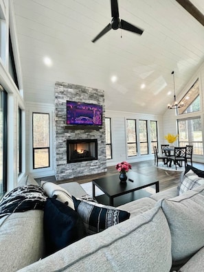 A spacious living room with a stone fireplace, a ceiling fan, and a TV mounted on the wall. - Little Darlin! Romantic Couples Cabin!!! Pet Friendly! (Hochatown)