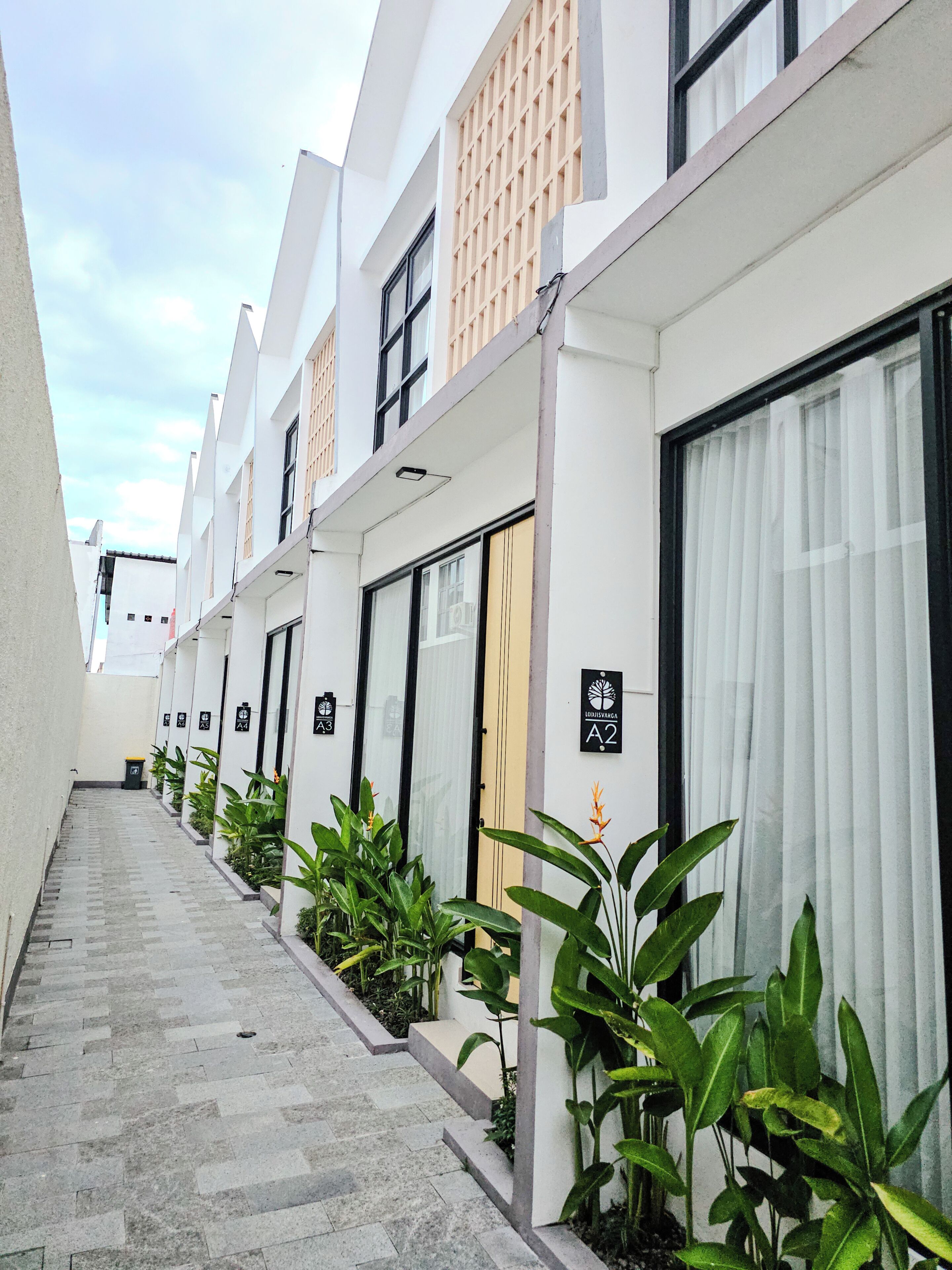 A row of modern, white buildings with numbered signs, a narrow paved walkway, and green plants along the sides.