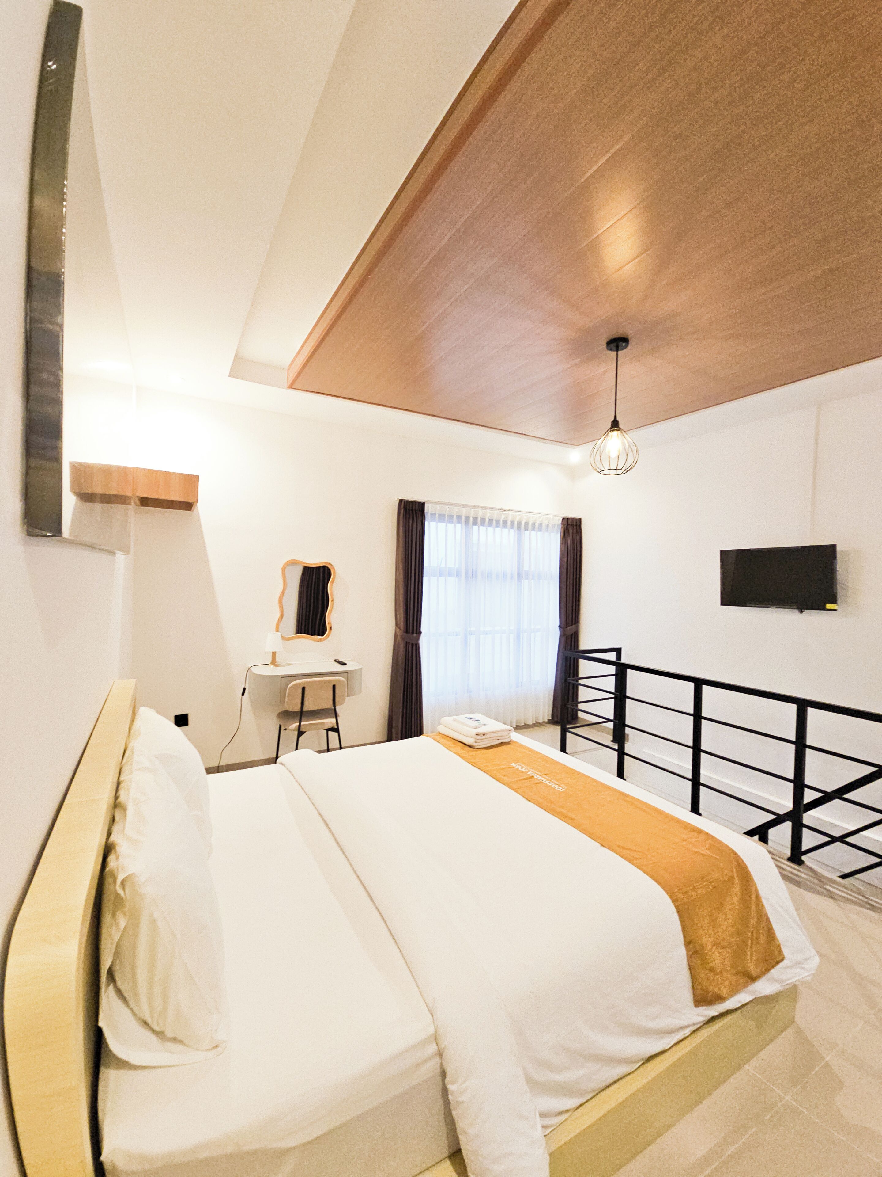 A hotel room with a bed, a desk, a chair, a window with curtains, a TV mounted on the wall, and a balcony railing.
