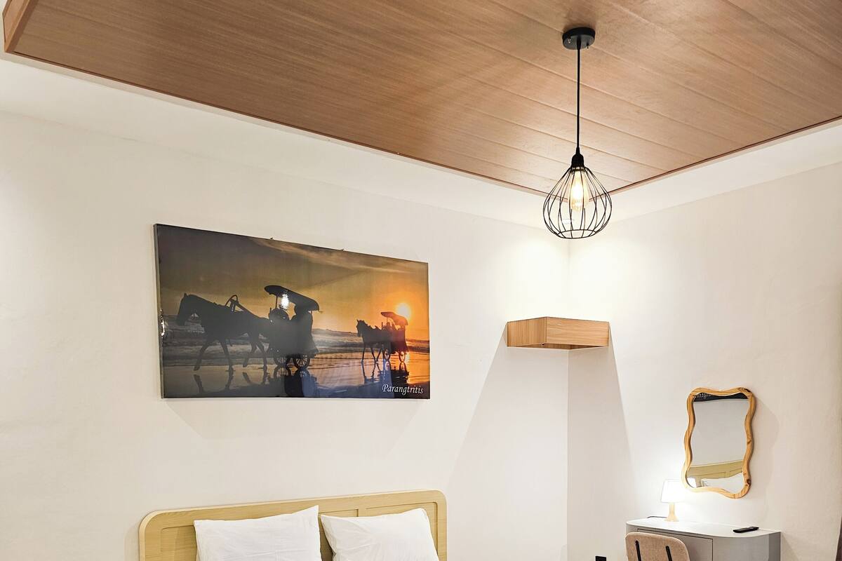 Habitació d'hotel moderna amb un llit gran, una taula de nit, un escriptori amb una cadira, un mirall fixat a la paret i un quadre amb una escena de capvespre.
