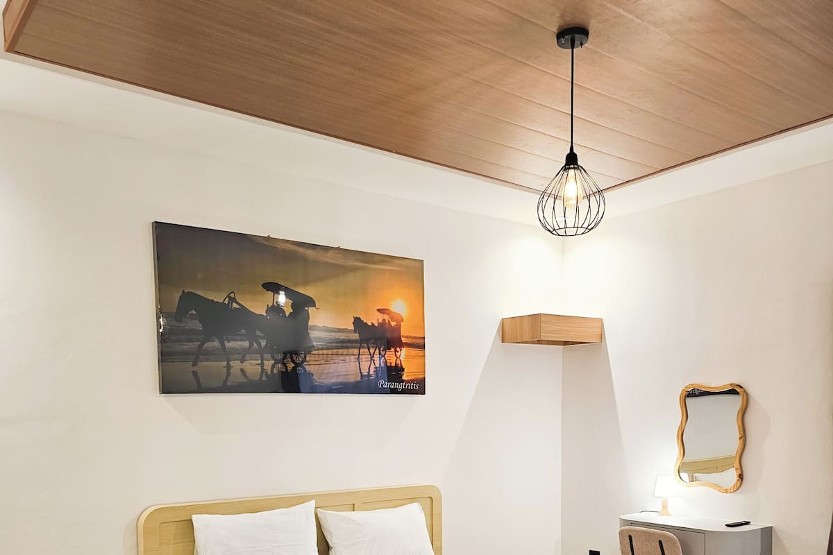 Una habitación de hotel moderna con una cama grande, una mesita de noche, un escritorio con una silla, un espejo en la pared y un cuadro de una escena de atardecer.