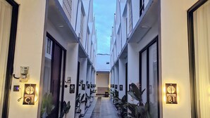 A corridor with a central walkway flanked by white buildings, each with a door and a sign indicating room numbers.