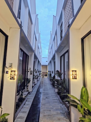 A corridor with a central walkway flanked by white buildings, each with a door and a sign indicating room numbers. - Lodji Svarga Premiere Villa (Sleman)